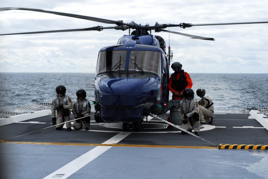 Den danska lynxhelikoptern surrad till däck med spännband. Under helikoptern syns gallret i vilket harpunen fäster. I orange dräkt syns den danska färdmekanikern. 
